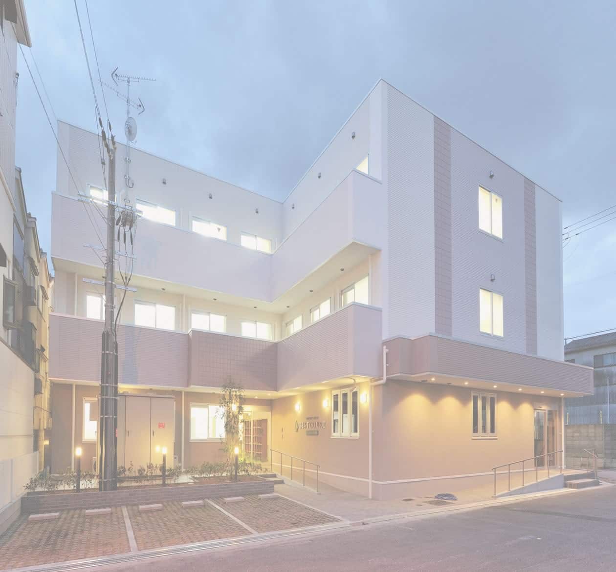 夕暮れに照らされた現代的な介護施設の建物外観。白とピンク系の外壁が特徴的。